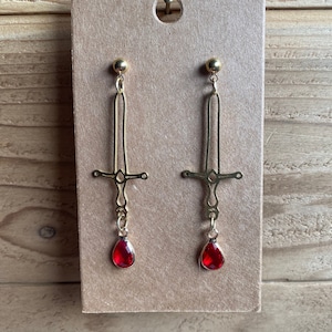 May include: Gold sword earrings with red teardrop-shaped crystals hanging from the bottom.