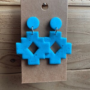 Pode incluir: Um par de brincos azul turquesa com um design de cruz geométrica. Os brincos são feitos de argila polimérica e têm uma textura salpicada.