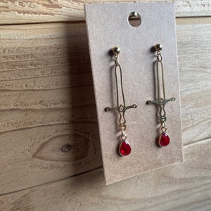 May include: A pair of gold earrings with a sword design and red teardrop-shaped crystals hanging from the bottom.