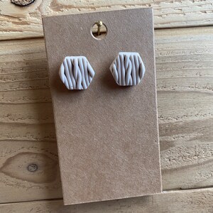 May include: A pair of white hexagonal stud earrings with a woven pattern. The earrings are displayed on a brown card.
