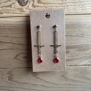 May include: A pair of gold sword earrings with red teardrop-shaped crystals hanging from the bottom.