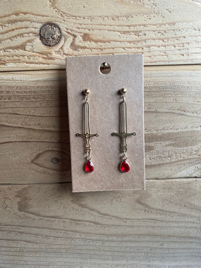 May include: A pair of gold sword earrings with red teardrop-shaped crystals hanging from the bottom.