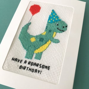 May include: A white card with a green dinosaur wearing a blue and yellow party hat and holding a red balloon. The card says "Have a roarsome birthday!"