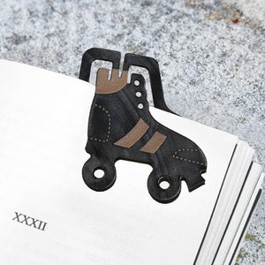 Puede incluir: Un marcapáginas de metal en forma de patín de ruedas negro y marrón. El marcapáginas tiene un diseño detallado. El marcapáginas está sujetando una página de un libro en la que está impreso el número XXXII.
