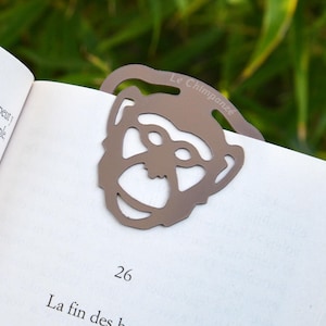 Peut inclure: Un marque-page en métal argenté en forme de visage de chimpanzé. Le marque-page porte le texte "Le Chimpanze" en haut. Le marque-page est sur un livre avec le numéro de page 26 visible.