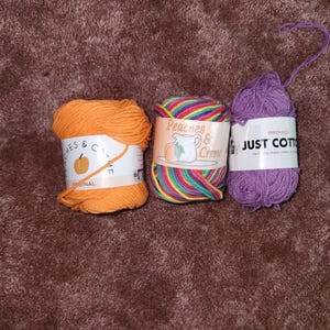 May include: Three balls of yarn in orange, rainbow, and purple. The orange yarn is labelled "Peaches & Creme Original". The rainbow yarn is labelled "Peaches & Creme". The purple yarn is labelled "Premier Just Cotton".