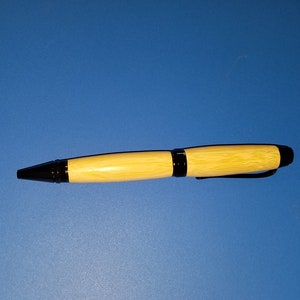 Puede incluir: Un bolígrafo de madera con un cuerpo de color claro y detalles en negro. El bolígrafo tiene una punta negra y un clip negro. El bolígrafo está sobre un fondo azul liso.