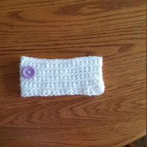 May include: A white, crocheted coffee cup sleeve with a lavender button fastening. The sleeve is rectangular and made of thick, textured yarn. The button is centred on one side of the sleeve.