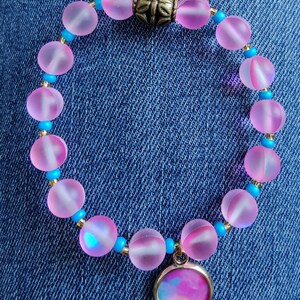 Könnte beinhalten: Ein rosa-blaues Perlenarmband mit einem goldenen Anhänger und einem rosa-blauen Glasanhänger. Das Armband besteht aus mattierten Glasperlen und hat einen goldenen Verschluss.