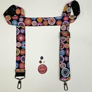 May include: A black crochet bag strap with a colorful circular pattern in shades of orange, pink, purple, and blue. The strap has metal hardware and a label that reads "@artisanosok".