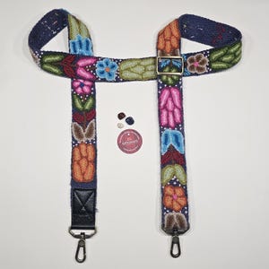 Könnte beinhalten: Ein farbenfroher, bestickter Taschenriemen mit marineblauem Hintergrund und floralen Mustern in Rosa, Orange, Grün und Blau. Der Riemen hat Metallbeschläge und einen Lederakzent. Ein kleines Logo und mehrere Perlen sind ebenfalls sichtbar.