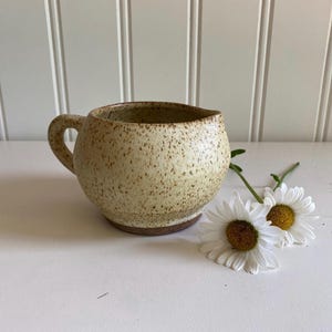 May include: A speckled ceramic creamer with a rounded body and a small handle. The creamer is beige with brown speckles and sits on a brown base. Two white daisies with yellow centres are next to the creamer.