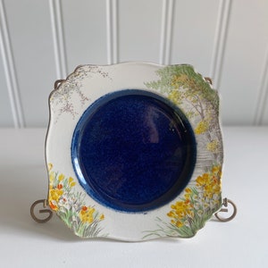 Puede incluir: Un plato pequeño de cerámica blanca con un borde festoneado. El plato tiene un centro azul y un diseño floral alrededor del borde. Las flores son amarillas, blancas y verdes.