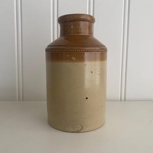 May include: A brown and cream ceramic jar with a rounded top and a slightly flared base. The jar has a brown band around the top and a brown glaze on the top half.