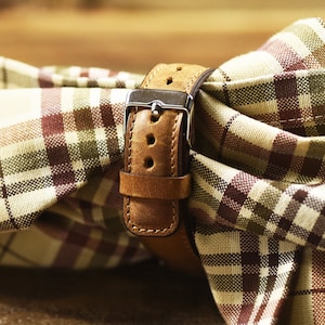 May include: A brown leather watch strap with a silver buckle. The strap is folded and placed on a plaid fabric.