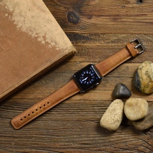 May include: A smartwatch with a black rectangular face and a tan leather strap featuring visible stitching and a silver-toned metal buckle. The watch display shows a circular analogue clock face with white numerals and hands against a black background.