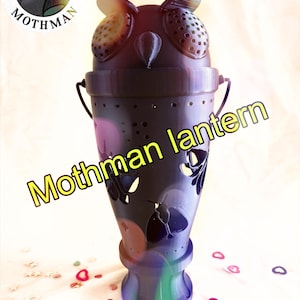 May include: A dark purple and green lantern shaped like a creature with large eyes and wings, featuring cutouts of moths. The text "I SUPPORT MOTHMAN" appears in a circular logo at the top left, and the words "Mothman lantern" are printed diagonally.