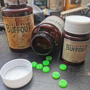 May include: Two brown bottles with white lids and labels that say "Original Buffout" and "50 Tablets". Green round pills are scattered on a dark surface. One bottle is lying on its side with the lid off.
