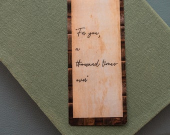 May include: A brown bookmark with a vintage newspaper design border. The bookmark has a quote on it that reads "For you, a thousand times over."