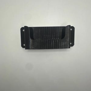 Puede incluir: Un soporte de pared negro de forma rectangular, impreso en 3D, con una abertura frontal en forma de U. El objeto cuenta con dos orificios de montaje circulares en cada pestaña lateral, diseñados para una fijación segura en superficies planas.