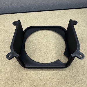May include: A black, 3D-printed mounting bracket featuring a central circular opening and four protruding tabs with pre-drilled holes for attachment. The structure has two vertical side walls and a flat base, constructed from a matte material.