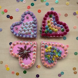 May include: Four pink felt hearts decorated with colorful sequins and beads. The hearts are arranged in a square shape.