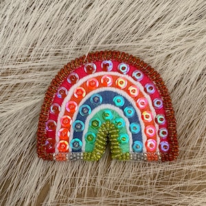 Könnte beinhalten: Ein gestickter Regenbogen-Patch mit Pailletten. Der Regenbogen ist aus rotem, orangefarbenem, gelbem, grünem, blauem und violettem Garn gefertigt. Die Pailletten sind schillernd und verleihen dem Patch Glanz.