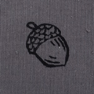 May include: A square, olive-green fabric patch with a black acorn illustration. The acorn is centered on the patch and has a detailed cap and a simple face. The patch has a scalloped edge and is set against a white, textured background.