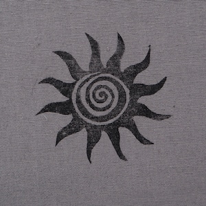 May include: A square, gray fabric patch with a black sun design. The sun has pointed rays and a spiral center. The fabric has a textured appearance and a white, scalloped edge.