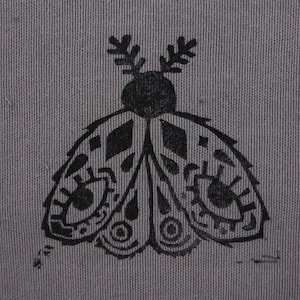 May include: A grey fabric square featuring a black moth design. The moth has detailed wing patterns and a round body. The fabric has a textured appearance and is bordered by a white, scalloped edge. The design is centred on the fabric.