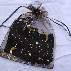 May include: A black drawstring bag with gold stars and crescent moons. The bag contains black thread, needles, and pins. The sheer outer layer of the bag is also decorated with gold stars and moons. The bag is tied with a black drawstring.
