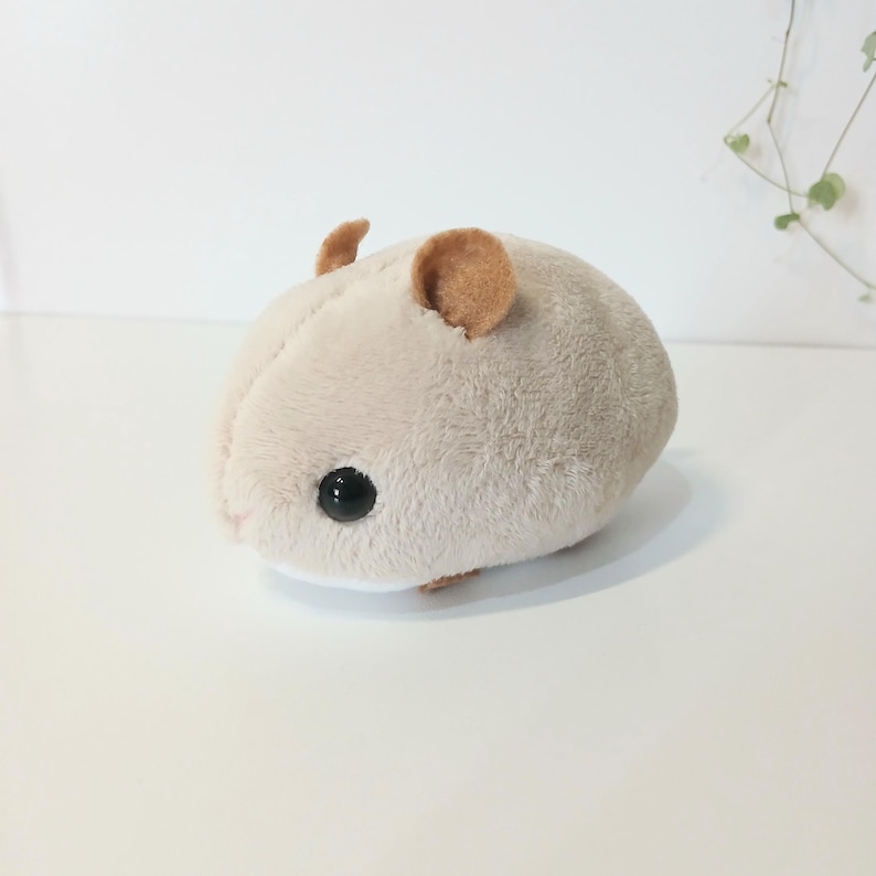 Pu&ograve; includere: Un peluche a forma di criceto di colore beige chiaro. Il giocattolo ha piccole orecchie arrotondate di colore marrone scuro e occhi neri a forma di perla. Il criceto &egrave; appoggiato su una superficie bianca.