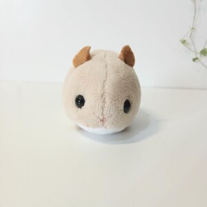 Pu&ograve; includere: Un peluche a forma di criceto di colore beige chiaro, con piccole orecchie marroni e occhi neri. Il giocattolo ha una pancia bianca e un piccolo naso rosa. Il criceto &egrave; rotondo e morbido.
