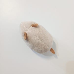 Pu&ograve; includere: Un peluche a forma di topo beige chiaro. Il giocattolo ha piccole orecchie marroni e una coda marrone. Il topo &egrave; appoggiato su una superficie bianca.