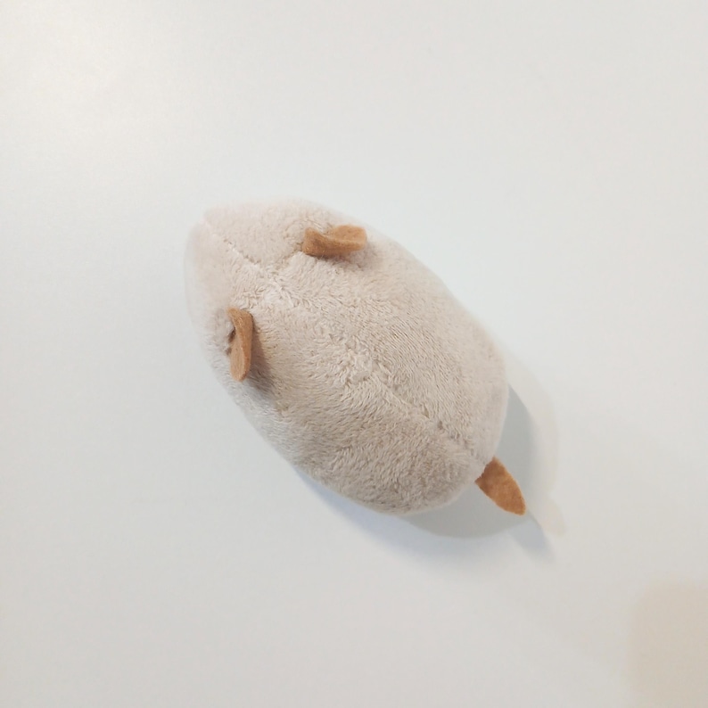 Pu&ograve; includere: Un peluche a forma di topo beige chiaro. Il giocattolo ha piccole orecchie marroni e una coda marrone. Il topo &egrave; appoggiato su una superficie bianca.