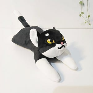 May include: A plush black and white cat toy with yellow eyes and a pink nose. The cat is lying down with its front paws extended. The tail is black with a white tip. The cat's ears are white.