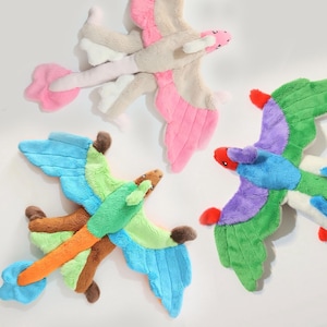 Peut inclure: Trois jouets dragons en peluche avec des textures douces et duveteuses. L'un est beige et rose, l'un est bleu, vert et orange, et l'autre est vert, violet et bleu. Chaque dragon présente des ailes déployées, une longue queue et des détails faciaux cousus.