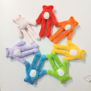 May include: A circular arrangement of seven plush frog toys in various colours including red, orange, yellow, green, blue, purple, and light pink. Each soft toy features a fuzzy texture, a white circular belly patch, and a simple stitched face.