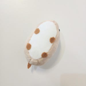 Pu&ograve; includere: Un giocattolo di peluche di forma ovale, nei toni del beige, bianco e marrone. Il giocattolo ha un centro bianco con quattro macchie marroni e un bordo beige. Una piccola coda marrone si estende da un'estremit&agrave;. Il giocattolo &egrave; progettato per assomigliare a un piccolo animale.