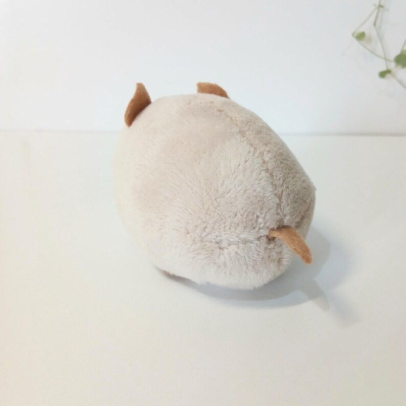 Pu&ograve; includere: Un peluche beige chiaro a forma di animale, forse un maiale, con piccole orecchie e coda marroni. Il giocattolo ha una consistenza morbida e soffice, su una superficie bianca. Un oggetto carino e coccoloso.