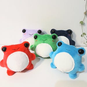 May include: A collection of five plush frog toys in red, green, blue, purple, and dark blue. Each frog features a round white belly, large black plastic eyes, and small embroidered mouth details. The soft toys are arranged in a group on a surface.