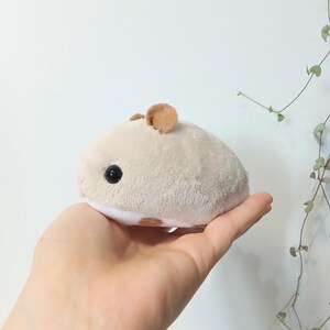 Pu&ograve; includere: Un peluche a forma di criceto di colore beige chiaro, con orecchie marroni e piccole macchie marroni. Il giocattolo ha un occhio nero ed &egrave; tenuto in una mano.