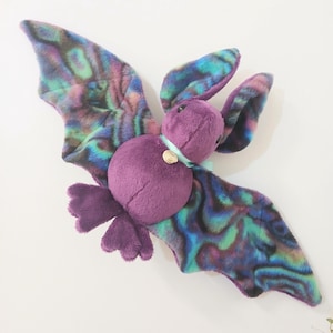 May include: A plush bat toy featuring a solid purple body and head with large, upright ears. The wings display a vibrant, iridescent pattern of swirling blue, green, and purple hues. A light blue ribbon with a small gold circular charm is tied around the neck.