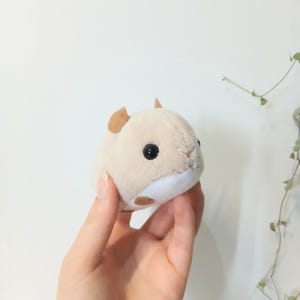 Pu&ograve; includere: Un peluche a forma di criceto beige e bianco, con piccole orecchie marroni e occhi neri. Il giocattolo di peluche &egrave; tenuto in mano su uno sfondo bianco. Il criceto ha un corpo rotondo e soffice, con un'espressione carina.