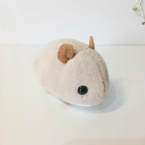 Pu&ograve; includere: Un peluche a forma di criceto beige chiaro con orecchie marroni e un occhio nero. Il peluche &egrave; morbido e rotondo, progettato per assomigliare a un criceto.