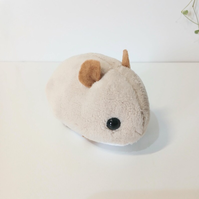 Pu&ograve; includere: Un peluche a forma di criceto beige chiaro con orecchie marroni e un occhio nero. Il peluche &egrave; morbido e rotondo, progettato per assomigliare a un criceto.