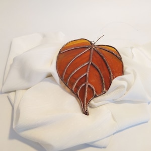 May include: A stained glass leaf ornament in a warm amber color with a copper wire frame. The leaf is resting on a white fabric surface.
