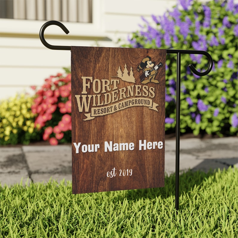 May include: A brown wooden garden flag with a cartoon mouse holding a rifle. The text "Fort Wilderness Resort & Campground" is in gold lettering. The text "Your Name Here" and "est 2019" are also on the flag.