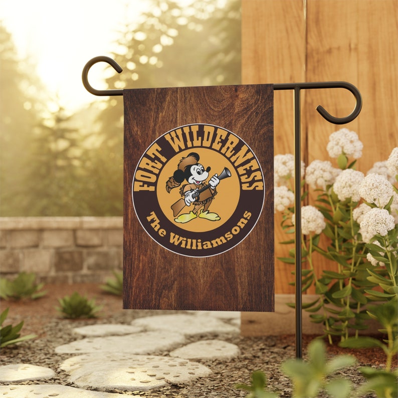 May include: A brown wooden garden flag with a yellow circle featuring a cartoon mouse wearing a cowboy hat and holding a rifle. The text "Fort Wilderness The Williamsons" is inside the circle.