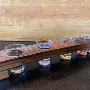 May include: A wooden tasting tray with five small clear glass shot glasses filled with different colored liquids. The tray is made of dark wood with a rustic finish.